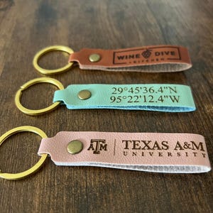 May include: Three leather keyrings with gold-coloured key rings. One is brown with "WINE DIVE KITCHEN" text, another is teal with coordinates, and the last is pink with "ATM TEXAS A&M UNIVERSITY" text.