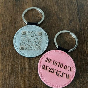 Puede incluir: Dos llaveros redondos. Uno es plateado con un c&oacute;digo QR, el otro es rosa con coordenadas: 29&deg;46'10.0"N 95&deg;23'47.1"W. Ambos tienen anillas plateadas y detalles en cuero negro. Los llaveros est&aacute;n sobre una superficie de madera.
