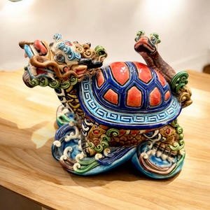May include: A ceramic sculpture of a mythical creature, a dragon-turtle hybrid. The sculpture features a red and blue shell, a dragon's head, and is set on a base with wave details. The colors include blue, red, green, and gold.