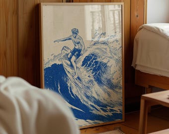May include: A framed print featuring a blue and white illustration of a surfer riding a large wave. The artwork depicts a person on a surfboard, with the wave cresting behind them. The frame is a light wood color.