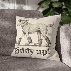 May include: A cream-colored throw pillow with a black and white illustration of a dog wearing a cowboy hat and boots. The text "Giddy Up!" is printed below the dog.