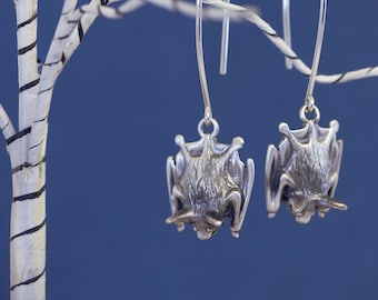 May include: Silver dangle earrings featuring a detailed bat design hanging from a silver wire. The earrings are hanging from a white branch against a blue background.