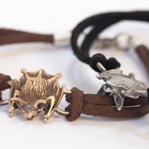 Könnte beinhalten: Zwei Lederarmbänder mit Fledermaus-Anhängern. Ein Armband ist aus braunem Leder, das andere aus schwarzem Leder. Die Fledermaus-Anhänger sind beide silbern und haben ein detailliertes Design.