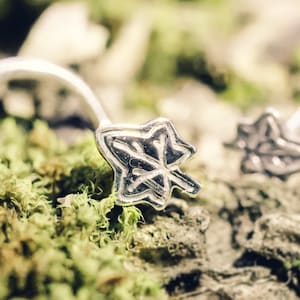 Puede incluir: Dos anillos de nariz de plata con un diseño de hoja. Los anillos están sobre una superficie musgosa.