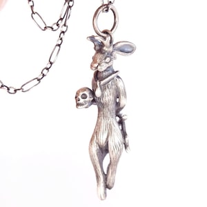 May include: A silver-toned pendant necklace featuring a stylized rabbit figure holding a skull. The rabbit wears a collar and has long ears. The pendant hangs from a delicate chain.