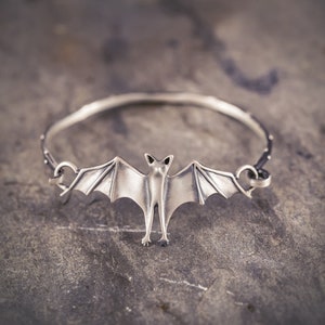 May include: A silver bangle bracelet with a bat charm. The bat is detailed with wings and a body.