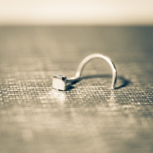 May include: A silver nose ring with a square-shaped stud. The ring is lying on a textured surface.