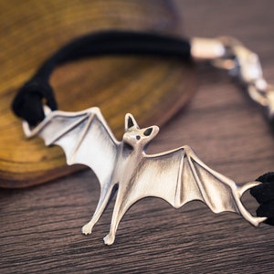 May include: A silver bat charm bracelet with a black leather cord. The bat charm is detailed with wings and a textured finish.