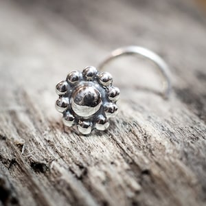 May include: A silver nose ring with a flower design. The flower has a round center and is surrounded by small, round petals.