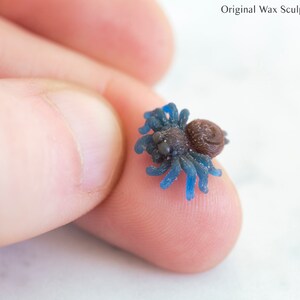 May include: A small blue wax sculpture of a spider with a brown body. The spider is being held by a finger.