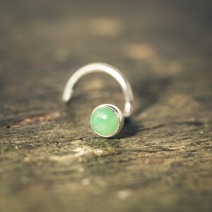May include: A silver nose ring with a green gemstone. The ring is curved and has a small, round stone.