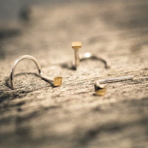 Op de afbeelding: Drie zilveren neusringen met gouden vierkante accenten. De ringen liggen op een houten oppervlak.