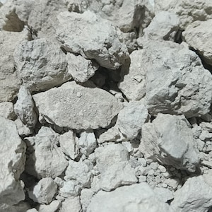 Pode incluir: Um monte de rochas cinza claro, de forma irregular. As rochas variam em tamanho, algumas parecem estar quebradas ou lascadas. A textura geral é áspera e irregular, sugerindo um material natural e não refinado. A imagem está bem iluminada.