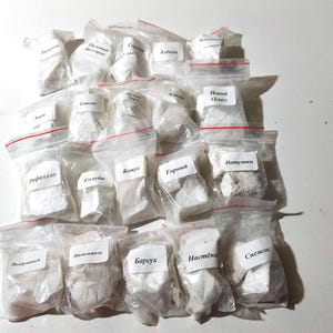 May include: A collection of small, clear plastic bags filled with white and off-white substances. Each bag is sealed with a red zip and has a small white label with handwritten text. The bags are arranged on a white surface.