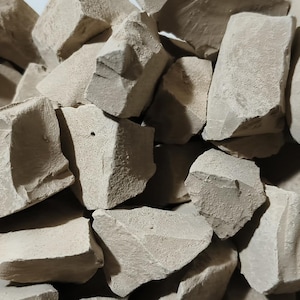 May include: A pile of irregularly shaped, light brown clay chunks. The pieces vary in size and have rough, textured surfaces. The clay appears to be natural and unprocessed, with a matte finish. The image is a close-up, showing the details of the clay.