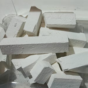 Puede incluir: Un recipiente de plástico transparente lleno de trozos de tiza blanca. La tiza tiene varias formas rectangulares, algunas rotas, y parece tener una superficie texturizada. El recipiente es parcialmente visible.