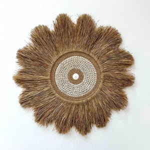 Op de afbeelding: Een ronde, geweven wandhanger met een sunburst-ontwerp. Het midden heeft een cirkel van kleine witte schelpen, omgeven door een geweven rand. De buitenrand heeft lange, bruine, stro-achtige vezels die naar buiten stralen.