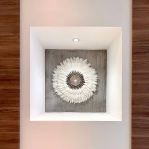 Puede incluir: Una obra de arte mural de plumas blancas con un diseño circular, con un centro marrón y dorado. La obra está montada sobre un tablero cuadrado gris y empotrada en un nicho de pared blanco. La pared está pintada de blanco y tiene paneles de madera.