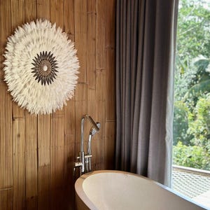 Puede incluir: Un adorno de pared circular de plumas blancas con un centro tejido está montado en una pared de bambú. El baño cuenta con un grifo cromado, una bañera curva y una cortina gris. La ventana ofrece una vista de exuberante vegetación.