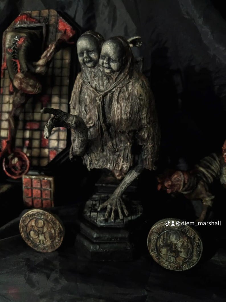 May include: A dark, textured sculpture of two conjoined figures with serene expressions, draped in weathered fabric. The sculpture has outstretched arms and is set on a hexagonal base. Two circular, aged coins are in the foreground.