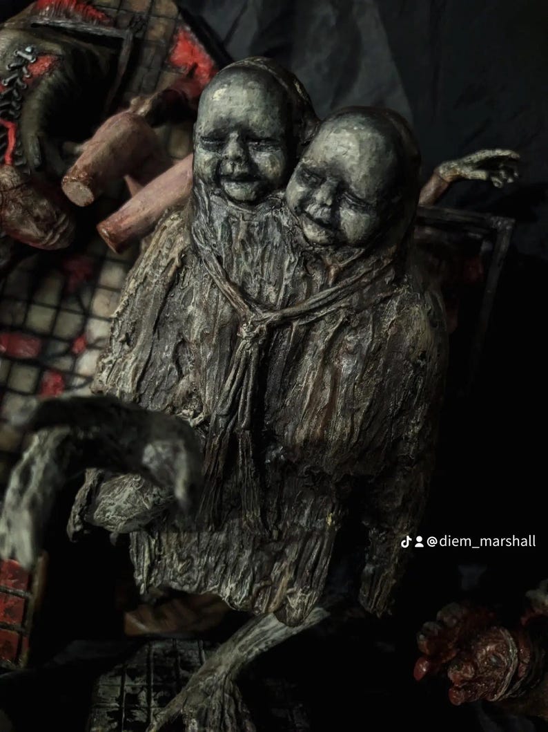 May include: A sculpture of two conjoined baby heads with closed eyes, attached to a textured, dark gray body. The sculpture has a distressed, aged appearance, with visible details and a somber expression. The overall theme is gothic and macabre.