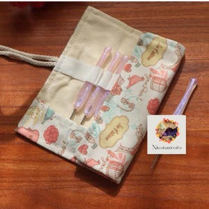 May include: A cream-colored crochet hook roll with a Parisian-themed pattern in pink, blue, and beige. The roll contains several pink crochet hooks. The roll is secured with a white strap and a beige drawstring.