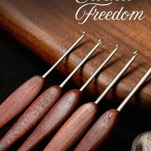 Peut inclure: Cinq crochets avec des pointes en métal argenté poli et des manches en bois brun foncé disposés en rangée. Le texte JIN LAN Crochet Freedom est imprimé en or et blanc en haut. Une petite pelote de fil blanc cassé est visible dans le coin.