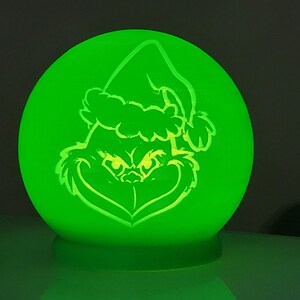 May include: A glowing green sphere featuring a cartoon outline of a character wearing a Santa hat. The character's face is visible, with a mischievous grin and furrowed brow. The light-up design is perfect for holiday decor.