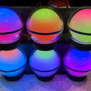 Puede incluir: Seis orbes de vidrio con una banda negra alrededor del medio, cada uno contiene un degradado de color diferente. Los colores son rosa, amarillo, verde, azul, morado y naranja.