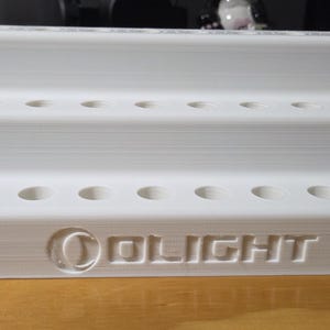 Könnte beinhalten: Ein weißer, mehrstufiger Präsentationsständer mit zahlreichen kreisförmigen Löchern zur Organisation von Gegenständen. Der Ständer hat drei Ebenen und das Wort "OLIGHT" ist auf der Vorderseite geprägt. Der Ständer besteht aus einem festen, weißen Material.