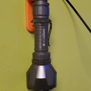 Peut inclure: Une lampe torche grise avec le texte "WARRIORX" et "T6" imprimé sur le côté est montée sur un support mural orange. La lampe torche est reliée à un cordon noir.