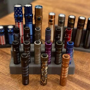 Op de afbeelding: Een verzameling van 20 zaklampen in verschillende kleuren en afwerkingen, waaronder zwart, zilver, goud, rood, blauw en paars. De zaklampen zijn in twee rijen gerangschikt op een grijze en zwarte displaystandaard.