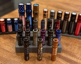 May include: A collection of 20 flashlights in various colors and finishes, including black, silver, gold, red, blue, and purple. The flashlights are arranged in two rows on a gray and black display stand.