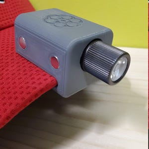 以下が含まれることがあります： 赤いボタンと黒い懐中電灯が取り付けられた、グレーの3Dプリントされた懐中電灯クリップ。クリップは赤い帽子に取り付けられています