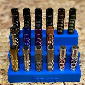 Könnte beinhalten: Eine Sammlung zylindrischer Metallobjekte in verschiedenen Farben, darunter Schwarz, Gold, Silber und Blau, die auf einem blauen, mehrstufigen Ständer ausgestellt sind. Die Objekte haben strukturierte Oberflächen und schwarze Oberteile. Der Ständer trägt den Text "5T EOS".