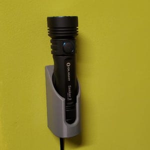 May include: A black Olight Seeker 2 flashlight is mounted on a gray wall mount. The flashlight is plugged into an outlet.