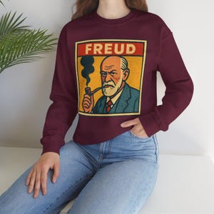 Może przedstawiać: Bordowa bluza z grafiką w stylu vintage przedstawiającą Zygmunta Freuda palącego fajkę, z napisem "FREUD" w czerwonym prostokącie. Ilustracja ma ż&oacute;łte tło i niebieski garnitur.