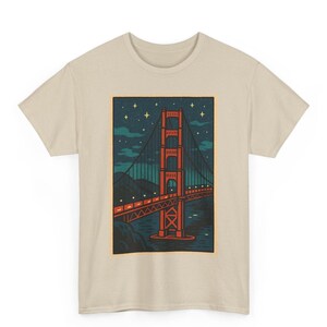 May include: Beige t-shirt featuring a graphic of the Golden Gate Bridge at night. The bridge is rendered in red with a dark blue sky filled with stars and clouds. The shirt is made of a soft, neutral-colored fabric.