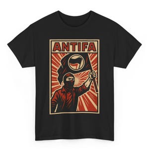 May include: Black t-shirt with a graphic design. The design includes the word "ANTIFA" in red, above an illustration of a person in a red jacket and black mask holding a flag with a circular symbol. The background has a radial design.