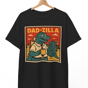Op de afbeelding: Zwart T-shirt met een retro-illustratie van een Godzilla-achtig wezen dat een kopje koffie vasthoudt. De tekst "DAD-ZILLA" staat boven de illustratie.