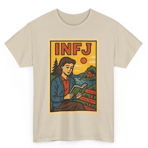 May include: Beige t-shirt featuring a retro-style graphic. The graphic depicts a woman reading a book, with the letters "INFJ" at the top. The illustration includes a landscape with a lake, trees, and a house, all in a vintage aesthetic.