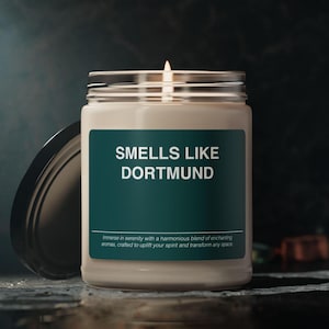 May include: A white candle in a glass jar with a green label that reads "SMELLS LIKE DORTMUND". The label also includes the text "Immerse in serenity with a harmonious blend of enchanting aromas, crafted to uplift your spirit and transform any space."