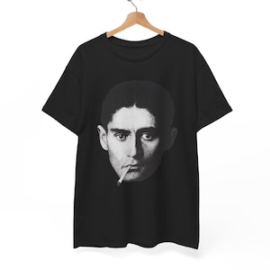 Może przedstawiać: Czarny t-shirt z czarno-białym portretem osoby z papierosem. Obraz jest wyśrodkowany. T-shirt wisi na drewnianym wieszaku.