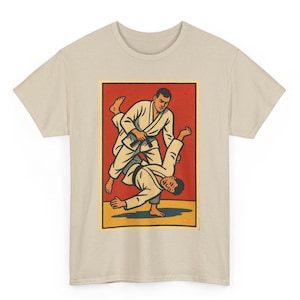 Könnte beinhalten: Ein beiges T-Shirt mit einer Grafik, die zwei Personen in weißen Kampfsportuniformen bei einem Judo-Wurf zeigt. Die Grafik ist vor einem roten und gelben Hintergrund mit schwarzen Umrissen und Details platziert.