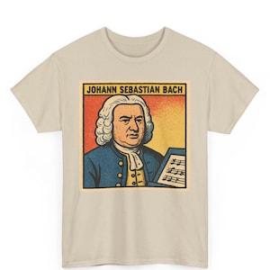 Pode incluir: T-shirt bege com um gráfico retrô de Johann Sebastian Bach. O design mostra Bach com um casaco azul, segurando partituras. O texto "JOHANN SEBASTIAN BACH" está no topo. Um design clássico para os amantes da música.