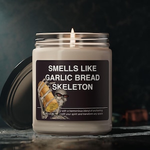 Op de afbeelding: Een witte kaars in een glazen pot met een zwart etiket waarop "SMELLS LIKE GARLIC BREAD SKELETON" staat. Het etiket toont een cartoon-skelet met een zonnebril dat een knoflookbrood vasthoudt.