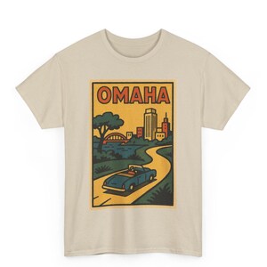 Op de afbeelding: Beige t-shirt met een retro-afbeelding van Omaha, Nebraska. Het ontwerp toont een cabriolet op een kronkelende weg, een stadsgezicht en het woord "OMAHA". De afbeelding is omlijst in een rechthoekige rand.