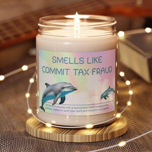 May include: A white candle in a glass jar with a pink, blue and yellow label that reads "Smells Like Commit Tax Fraud". The label also features a cartoon dolphin.