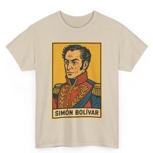 May include: Beige t-shirt featuring a vintage-style portrait of Simón Bolívar within a yellow rectangle. The portrait is detailed with red, blue, and gold accents. The name "SIMÓN BOLÍVAR" is printed in a bold font at the bottom of the rectangle.