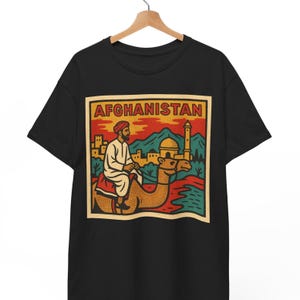 May include: Black t-shirt with a retro illustration of a person on a camel in Afghanistan. The design, framed with the word "AFGHANISTAN", uses red, yellow, green, and brown. The image evokes a sense of travel and cultural heritage.